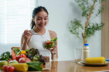 Happy young asian woman eating a healthy salad for breakfast or lunch, sitting at the table in a...