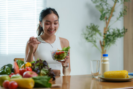 Happy young asian woman eating a healthy salad for breakfast or lunch, sitting at the table in a bright modern kitchen, enjoying fresh organic vegetables and healthy lifestyle