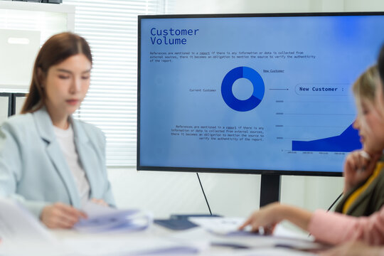 A business meeting scene featuring a woman reviewing documents while data on customer value is displayed on a screen.