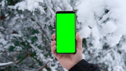 Young man's hand holding smartphone with green screen against snowy forest background. Vertical mockup of mobile phone. Man using phone with blank green display background. Smartphone with chroma key