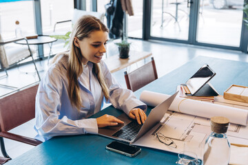 Female architect working on laptop with blueprints in modern office