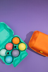 Vertical image in top view of colorful eggs in a blue painted basket next to an orange one on a purple background. Easter painted eggs