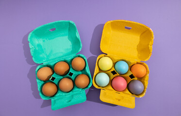 Top view of blue and orange egg cartons with colored eggs in orange basket and unpainted eggs in blue basket on a purple background. Easter decoration