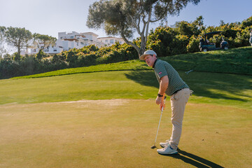 A golfer preparing to putt on a sunny golf course with greenery, trees, modern buildings, and a...
