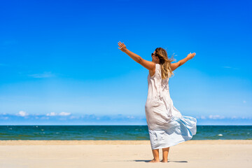 Beautiful middle aged woman in dress fluttering on wind standing with arms raised up on white sandy...