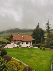 A small house with a red roof sits on a lush green lawn. The house is surrounded by trees and bushes, giving it a peaceful and serene atmosphere