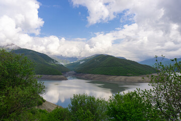 Reservoir on the river between two hills. Steep rocky banks. Spring green grass, bushes, trees. Mountains, hills and cloudy sky in the background