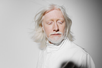 Man with white hair and beard, wearing a white outfit, against a plain light gray background,...