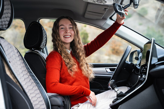 A female driver following safe ride rules, looking at the camera and smiling. A young friendly woman adjusting the rearview mirror in car before driving. Positive teenage girl in the driver's seat.