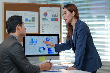 Businesswoman explaining financial charts on a computer to a male colleague in a modern office, engaging in a discussion about data analysis and corporate strategy