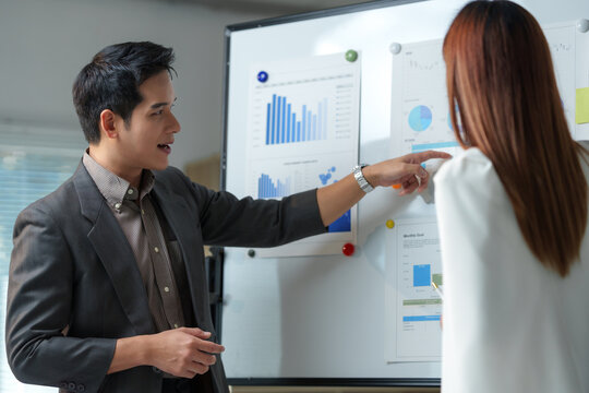 Businessman pointing at charts on whiteboard while giving a presentation to a female colleague in the office, discussing company performance, sales, and marketing strategy