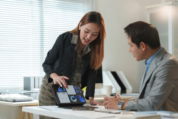 Asian businesswoman pointing at tablet screen showing financial charts and graphs to businessman colleague while working together on new project in modern office