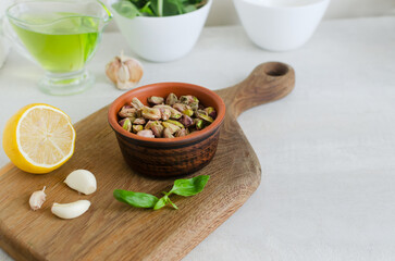 Peeled roasted pistachios in a ceramic bowl on a wooden board with basil, lemon and garlic. The process of making pistachio pesto. Horizontal orientation. Selective focus. Copy space
