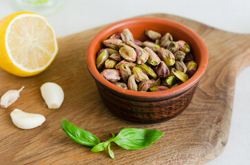 Peeled roasted pistachios in a ceramic bowl on a wooden board with basil, lemon and garlic. The process of making pistachio pesto. Horizontal orientation. Selective focus.