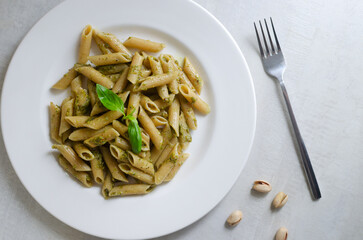 Penne pasta with pistachio pesto with a sprig of basil in a white plate with a fork on the table. Italian cuisine concept. Horizontal orientation. Selective focus. Top view