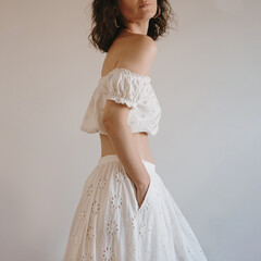 Pretty woman with loose curls in white off-shoulder dress in neutral setting. Relaxed pose,...