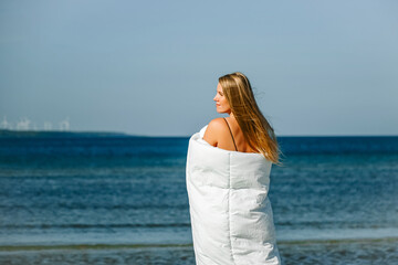 Obraz premium woman in white blanket walking by the sea on sandy beach. 