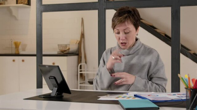 Confident middle aged female freelancer with hearing loss communicating with sign language with business colleague online, discussing project while making video call using digital tablet indoors..