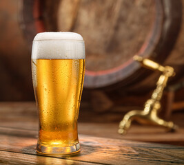 Glass of chilled beer on old wooden table and wooden beer cask and tap at the background.