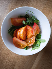 Fresh sliced tomatoes resting on lamb's lettuce, nestled in white ceramic bowl atop rustic wooden surface