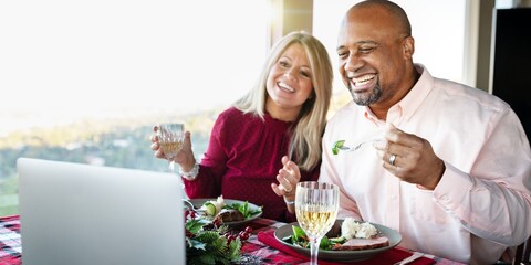 Smiling couple enjoying meal, holding wine glasses, sitting at table. They are engaged in a video call, sharing a joyful moment together. Couple having dinner and video call on laptop. Virtual dinner.