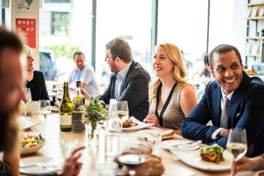 Group of people dining at a restaurant, enjoying food and conversation. Diverse group, mixed genders, smiling and socializing at a table with drinks and meals. Friends at festive lunch with wine.