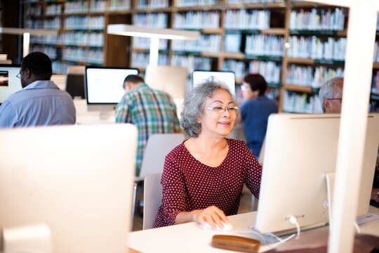 Elderly woman using a computer in a library. Focused senior working on a desktop. Learning and technology for older adults in a public space background with copy space. Elderly and technology concept.