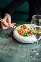 A close-up of a woman's hand holding chopsticks over a vibrant bowl of food, featuring vegetables and meat, with a glass of white wine beside it. The setting has a stylish, rustic ambiance.