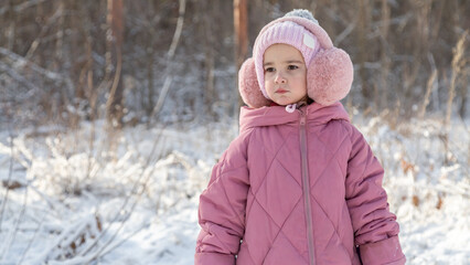 Cheerful positive child girl wearing a outerwear smiling walking through a snowy countryside forest