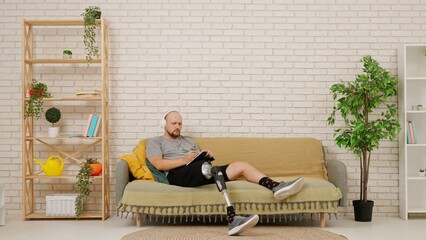 A man with a prosthetic leg sits on a sofa in a cozy living room, listening to music with headphones and writing down ideas in a notebook.
