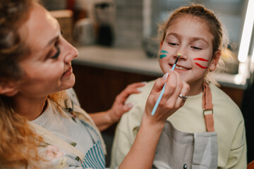 Mother painting daughter's face with colorful stripes in kitchen