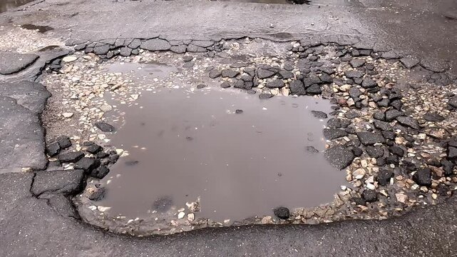 Rain is falling on a damaged asphalt road creating a puddle, showcasing the effects of weather and time on urban infrastructure during a winter day, pulling out shot