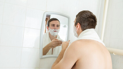 Young man with naked torso shaving his beard with foam looking at his reflection in mirror at...