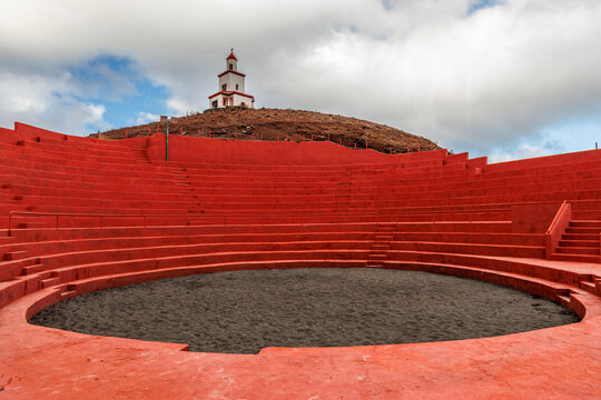 Vibrant red Canarian wrestling arena in El Hierro, Spain, under a dramatic sky. The traditional venue contrasts with the ocean and hills, evoking heritage and culture.