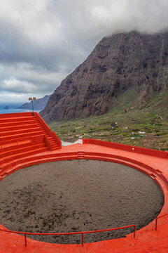 Vibrant red Canarian wrestling arena in El Hierro, Spain, under a dramatic sky. The traditional venue contrasts with the ocean and hills, evoking heritage and culture.