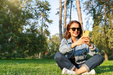 Smiling woman sits on green grass in lotus pose and texts on her phone.