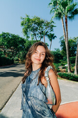 A smiling woman in a blue flowing dress and metallic bag poses on a sunny day surrounded by lush...