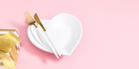 White heart shaped plate with fork and knife, beautiful gift with gold ribbon on pastel pink background to celebrate Valentine's Day, romantic dinner or wedding anniversary. Banner.