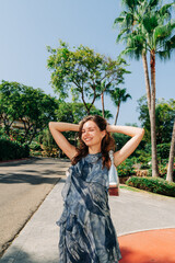 A carefree woman in a stylish dress smiles and stretches her arms above her head, enjoying the outdoors with a bright blue sky and greenery