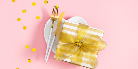 Top view of heart shaped plate with gift with ribbon and golden cutlery in center of composition on pink table background. Banner.