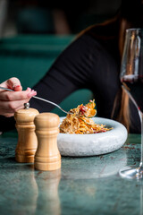 A close-up of a hand holding a fork with pasta, accompanied by a glass of red wine and wooden salt and pepper shakers on a stylish table. The setting is elegant and inviting.