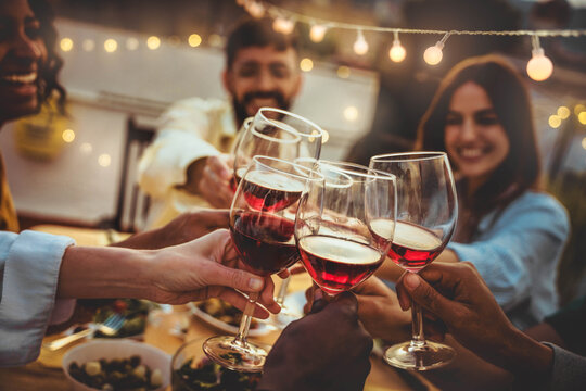 People toasting red wine glasses on rooftop dinner party - Happy friends eating meat and drinking wineglass at restaurant patio - Food and beverage lifestyle concept with guys and girls dining outdoor