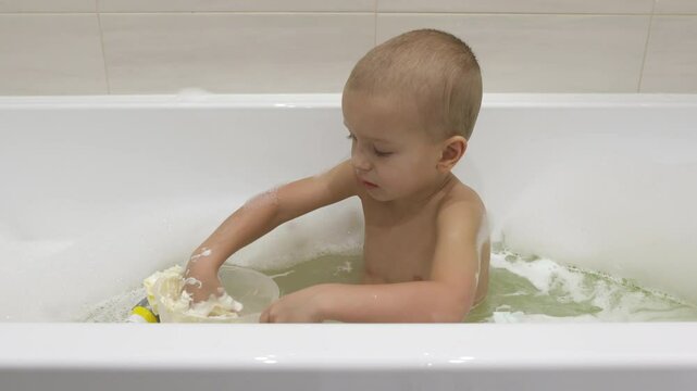 young boy is happily playing with a cup of bubbles in his bath, looking down and intently stirring the bubbles with his hand.