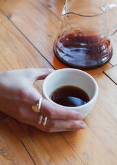 Young caucasian freelancer woman hold white cup with black filter coffee in glass kettle server. Barista on cafeteria giving mug to customer. alternative brewing, specialty, v60. background light wood