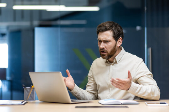 Frustrated and sad young businessman man sitting at desk in office and talking on video call on laptop gesturing with hands