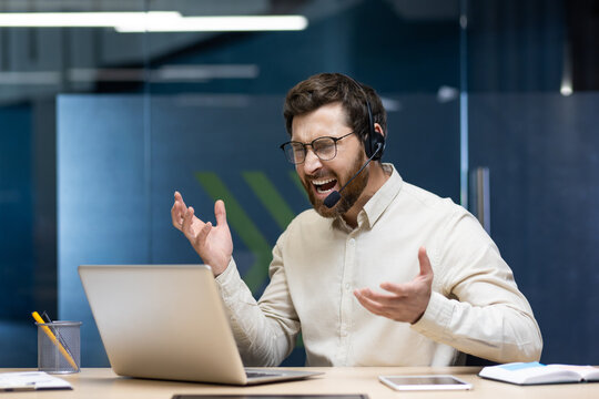 Young angry man in headset working in office on notebook, sitting at desk and emotionally shouting and arguing
