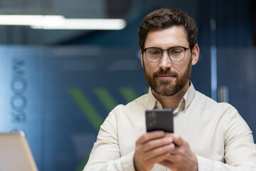 Close-up photo of a smiling young man in a shirt and glasses who is in the office and using a...