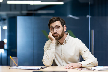 Portrait of a tired young man, an office worker sitting bored at the desk in the office and looking at the camera