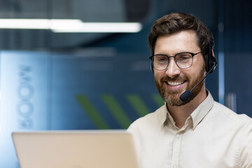 Close-up photo of a young smiling and self-confident man wearing a headset working at a laptop in the office