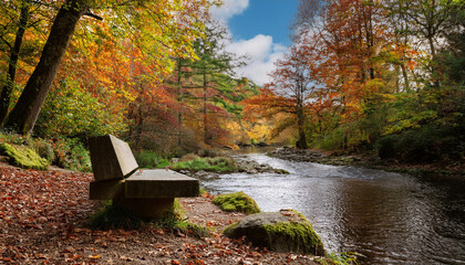 Obraz premium A concrete bench overlooks a peaceful river surrounded by trees displaying vibrant autumn foliage in orange and green hues.